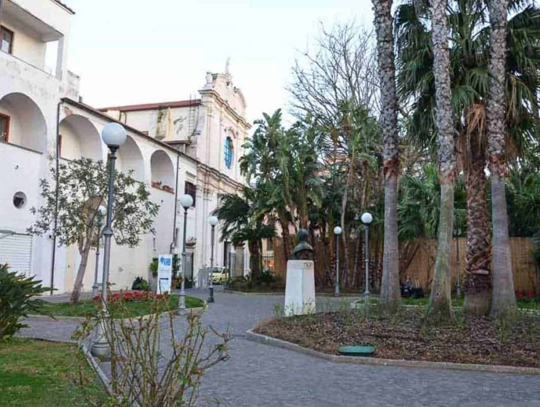 Vecchio edificio storico con giardino urbano e palme, atmosfera tranquilla e rilassante, ideale per passeggiate e relax, decorazioni eleganti con lampioni e statue, ambientazione suggestiva, perfetta per appunti sul blog di viaggi o cultura italiana.