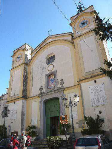 Palazzo storico con dettagli barocchi e chiesa fiancheggiata da alberi, ingresso principale con portale verde e orologio sulla facciata, in un tipico scenario urbano italiano.