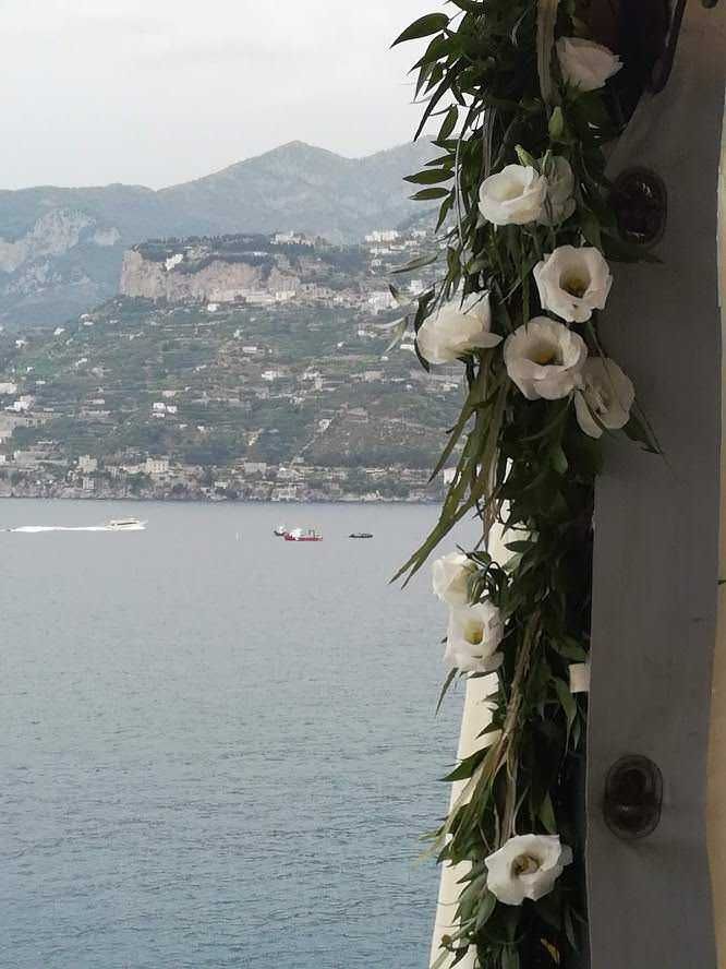 Vela decorata con fiori bianchi lungo il mare con paesaggio montano sullo sfondo, atmosfera romantica e tranquilla, ideale per eventi o momenti speciali in costiera.
