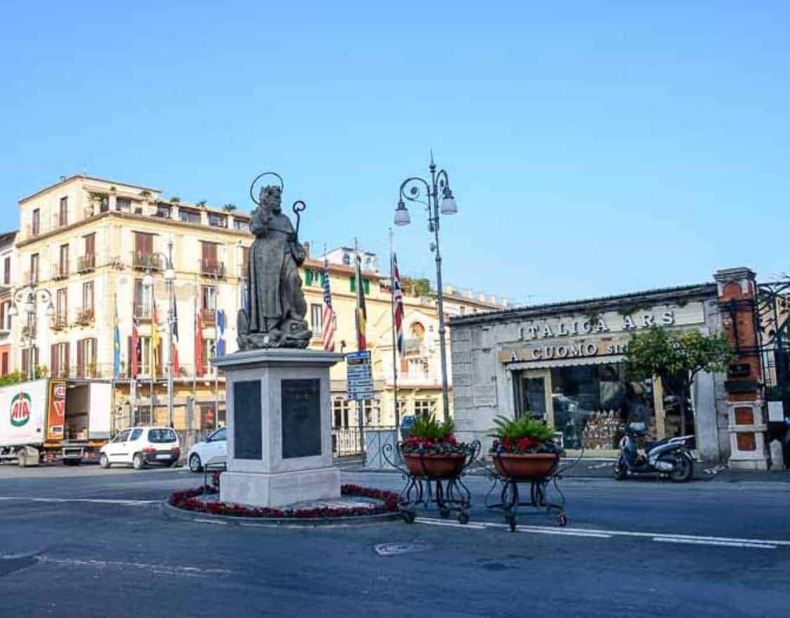 Statua di un santo in centro a Napoli, Italia, con negozi e auto sullo sfondo, simbolo di arte e cultura italiana.