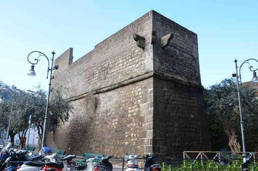 Antica torre di pietra con archi e lampioni in stile classico, situata al centro di una piazza storica, con scooter parcheggiati e alberi intorno, rappresenta un elemento storico e architettonico di rilievo.