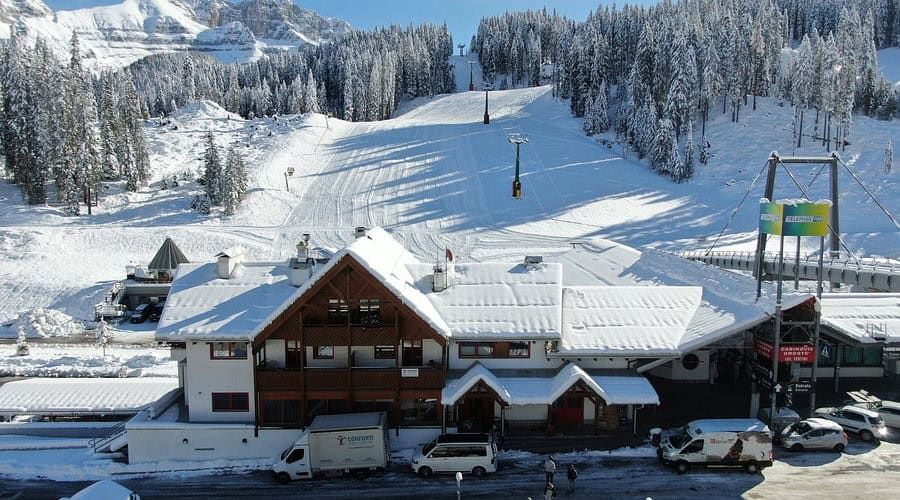 Immagine di un hotel a Madonna di Campiglio circondato da neve e boschi, con impianti di risalita e veicoli parcheggiati, ideale per chi cerca relax tra neve perenne e laghi azzurri.