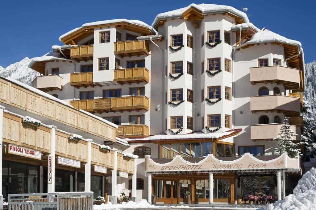 Hotel in Madonna di Campiglio con architettura alpina, situato tra neve perenne e laghi cristallini, ideale per vacanze invernali. Struttura moderna con balconi in legno e servizi di qualità.