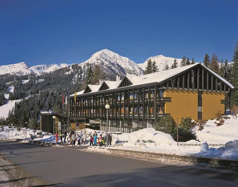 Hotel moderno immerso nella neve con montagne e laghi azzurri sullo sfondo, ideale per soggiorni invernali e relax tra natura e paesaggi mozzafiato.