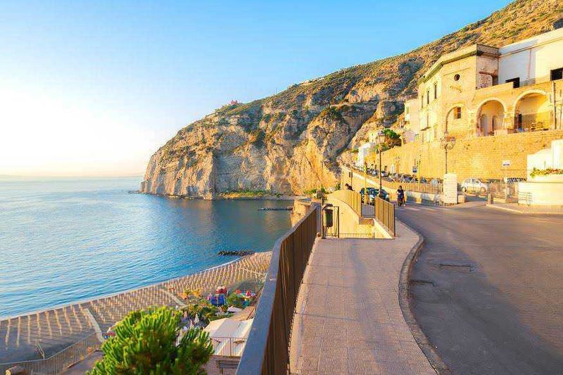 Quali sono le migliori spiagge di Sorrento di sabbia vulcanica e ciottoli? 7 Spiaggia di Amalfi con vista sul mare e scogliera alpina, passeggiata panoramica sulla strada costiera, edifici storici e atmosfera mediterranea al tramonto.