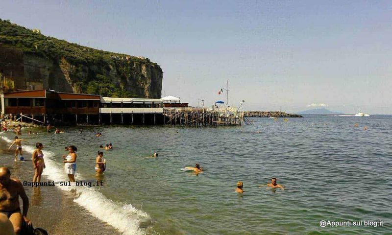 Quali sono le migliori spiagge di Sorrento di sabbia vulcanica e ciottoli? 3 Spiaggia affollata con acqua cristallina, stabilimenti balneari e scogliera sullo sfondo, perfetta per relax estivi e attività acquatiche nel mare italiano.