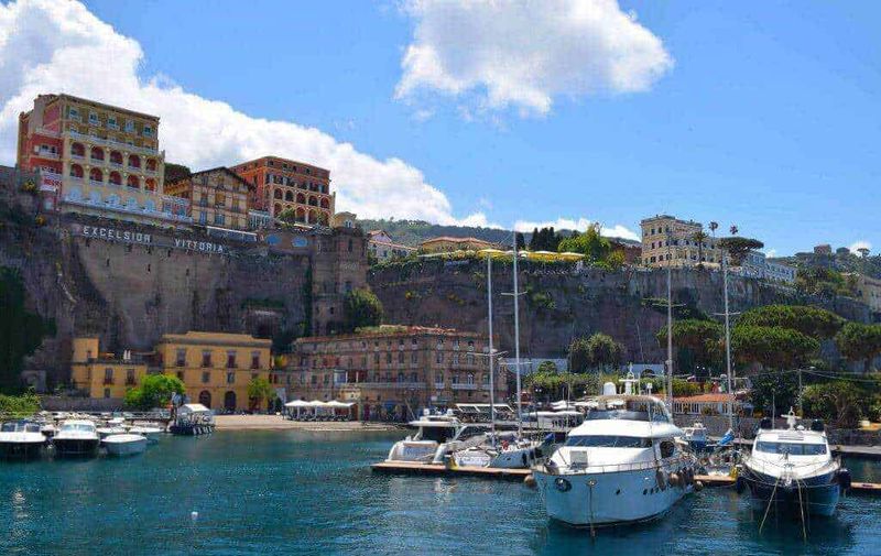 Quali sono le migliori spiagge di Sorrento di sabbia vulcanica e ciottoli? 2 Visto per prima cosa: Un porto nel colorato quartiere di Posillipo a Napoli, Italia, con yachts in mare e storiche abitazioni su scogliera, sotto un cielo azzurro con nuvole.