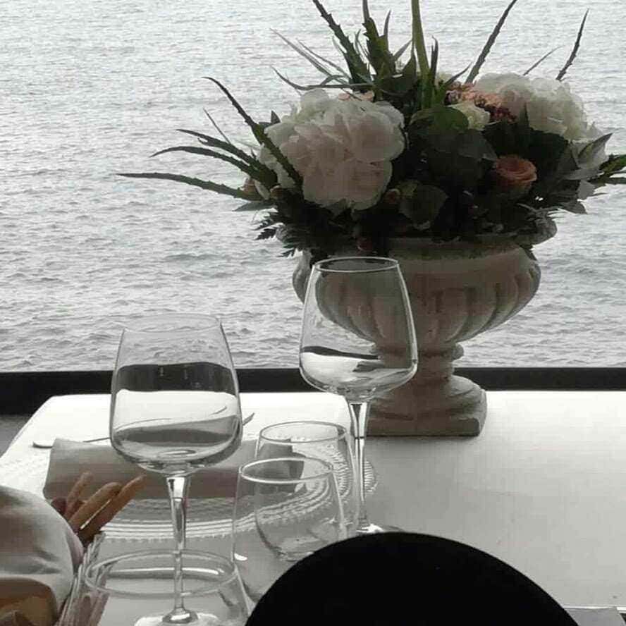 Significato colori rose: Significato dei colori delle rose: Fioriere di fiori bianche e rosa sulla tavola con bicchieri di vino e acqua, vista sul mare, atmosfera elegante e romantica, perfetta per un pranzo o una cena raffinata.
