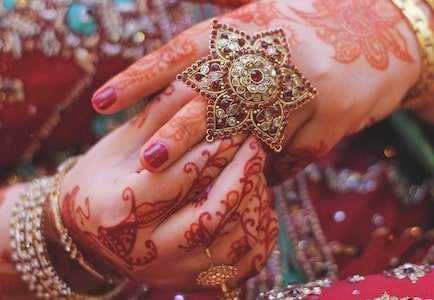 Anello d'oro tradizionale con pietre preziose, indossato su mani decorate con mehndi, simbolo di matrimonio e cultura indiana, circondate da abiti ricchi di dettagli e gioielli.