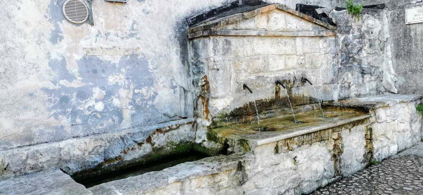 Fonte storica in pietra con fontana pubblica, acque che scorrono dai tubi di ferro, ambientazione urbana antica, caratteristiche di architettura tradizionale italiana, su muro di pietra.