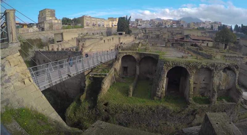 Ercolano: come arrivare e visita alle 15 antiche rovine romane del 79 dC 5 Antica area archeologica con rovine romane e struttura moderna di passerella in metallo, immersa in un contesto urbano, perfetta per appunti sulla storia e l'archeologia.