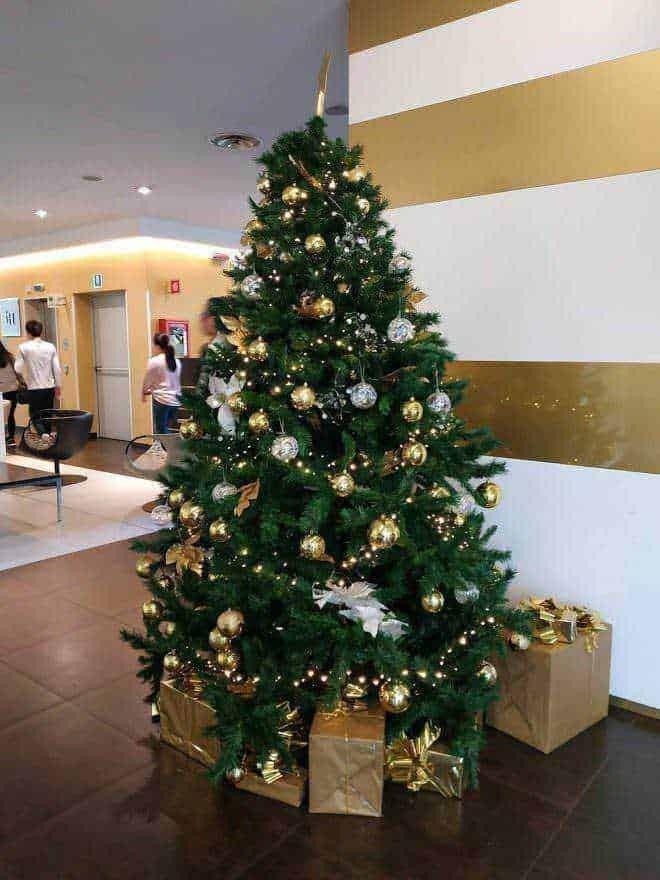 Alto testo: Albero di Natale decorato con sfere dorate e argento, addobbi natalizi, luci e regali sotto l'albero in un ambiente luminoso e accogliente.