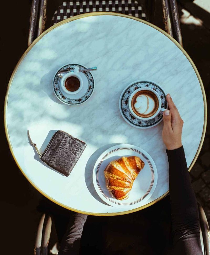 Tour di Napoli dal centro storico al lungomare Ovest 4 Una tazza di cappuccino con latte decorato, una tazzina di caffè, un croissant dorato e una borsetta su un tavolo in marmo di un bar elegante, perfetto per appunti sul blog.