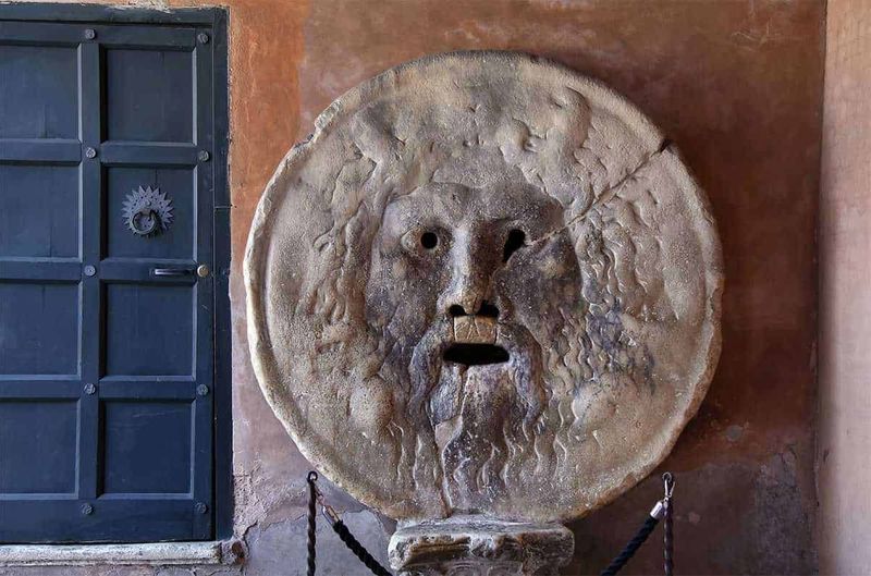 Roma passeggiando lungo il Tevere per 3 km, 14 curiosità 4 Antico monumento romano con testa di Medusa in pietra, decorazione storica su parete in mattoni rossi, adiacente a porta in legno blu, atmosfera storica e artistica.