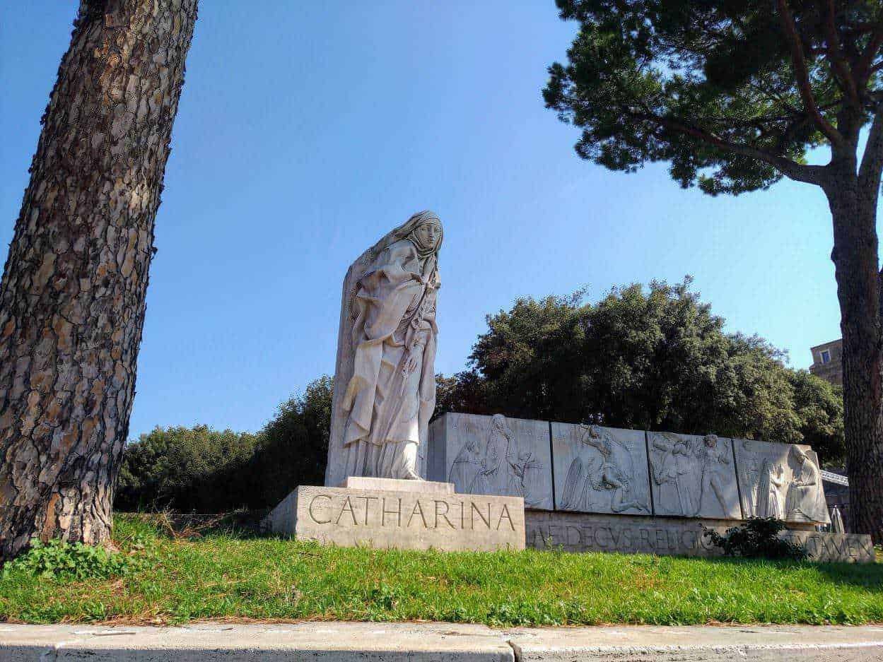 Statua di Santa Caterina in marmo con sfondo di alberi e cielo azzurro, monumento storico situato in un parco cittadino, omaggio alla santa con dettagli scolpiti e iscrizione sulla base.