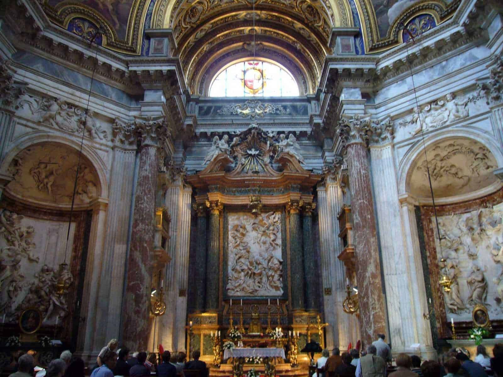 Maestoso interno barocco con decorazioni dorate, colonne in marmo e soffitto affrescato, chiesa storica con altare centrale, affluenza di fedeli, visitatori e dettagli artistici.