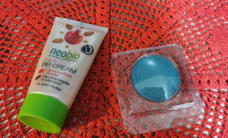 4 regole trucco antietà con BB, ombretto e matita per sopracciglia 2 Trucco antietà: Neobio BB Cream 7 in 1 con estratti di melograno e mandorle per un incarnato uniforme e idratato, posizionata su un tovagliolo rosso ricamato, ideale per la cura quotidiana della pelle.