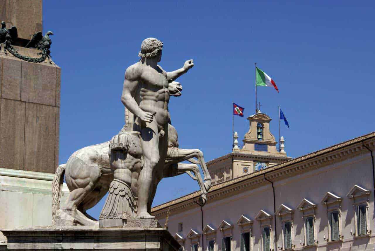 Statua di un mitologico centauro con sfondo edificio storico e bandiere italiane e europee, simbolo di cultura e storia italiana, su cielo sereno.