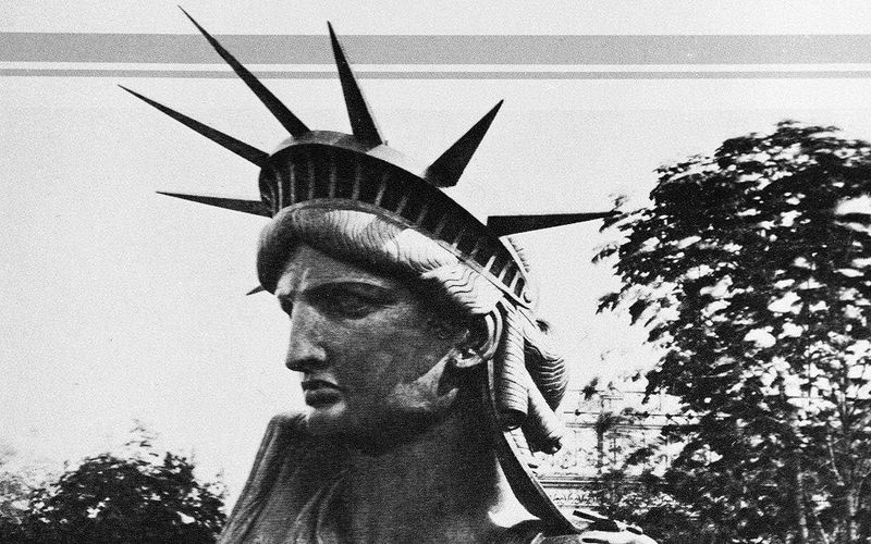 10 cose che non sapevi sulla Statua della Libertà 104 Statua della Libertà in stile vintage, foto in bianco e nero, simbolo di libertà e democrazia, raffigurante una donna con diadema a raggi. Scena urbana con alberi sullo sfondo, atmosfera retrò.