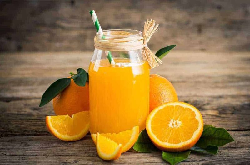 5 succhi di frutta fatti in casa per diabetici 3 Succo di arancia naturale in un barattolo di vetro con cannuccia verde, accompagnato da arance fresche e foglie verdi, sul tavolo di legno rustico, ideale come esempio di salutare bevanda energetica.