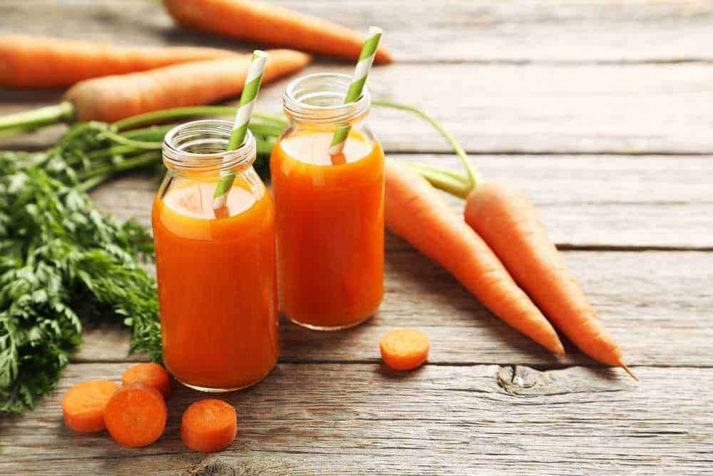 Vasetti di succo di carota fresco con bastoncini in una bottiglia di vetro, accanto a carote e foglie di prezzemolo su tavolo di legno naturale.