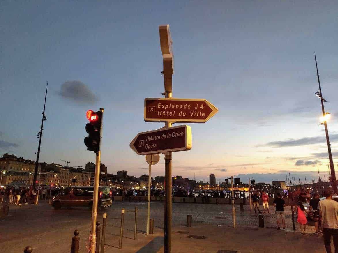 Palo con segnali stradali di indicazione a Marsiglia al tramonto, con il porto e le persone che passeggiano sul lungomare. Scena urbana vivace di sera con cielo al crepuscolo, ideale per blog di viaggio e turismo.