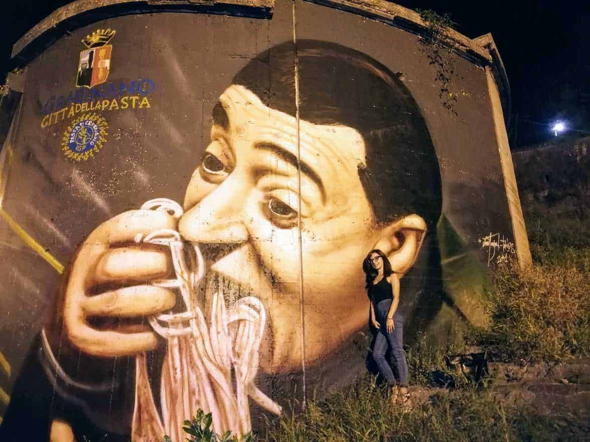 Vignetta di graffiti artistico rappresentante un uomo che sorseggia uno spaghetti, situato sul muro di una città italiana, con una donna in piedi accanto, sotto un cielo notturno.