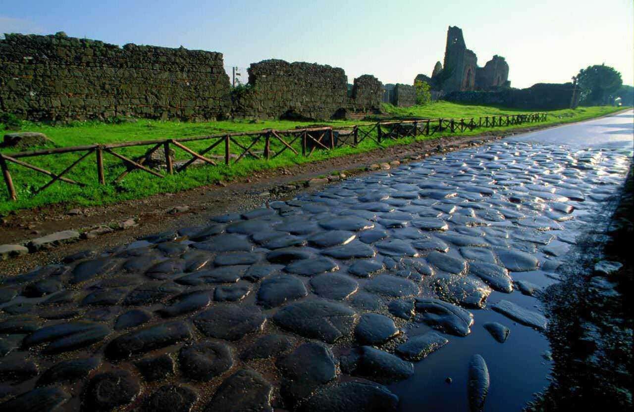 Antico camino di pietra con rovine romane sullo sfondo, sotto un cielo blu, immerso in un paesaggio verde naturale, ideale per esplorare storia e natura.