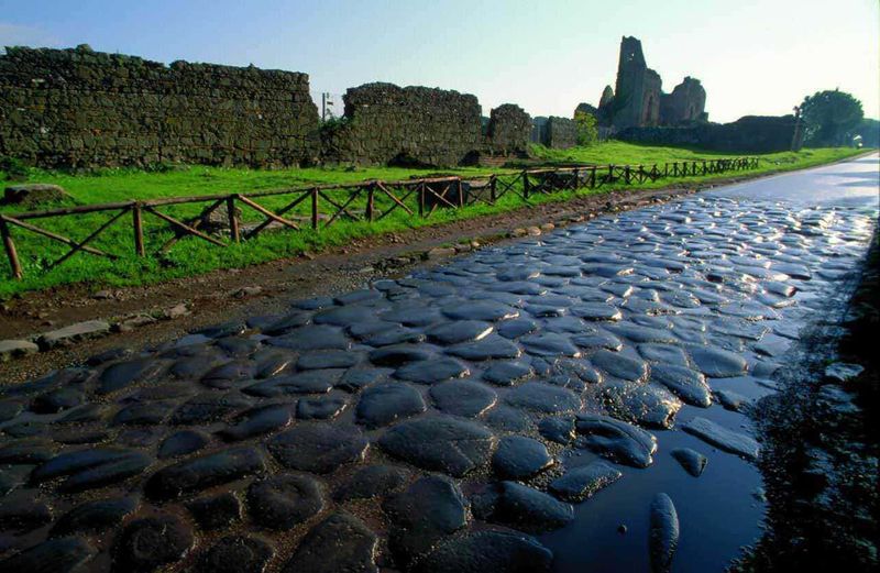 Via Appia Antica in bici sulla "Regina delle strade" a Roma 3 Antico camino di pietra con rovine romane sullo sfondo, sotto un cielo blu, immerso in un paesaggio verde naturale, ideale per esplorare storia e natura.