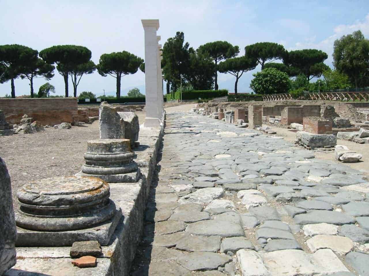 Antico sito archeologico di Roma antica con pavimentazione in pietra e resti di colonne, immerso nel verde di pino marittimo e cipressi, esempio di archeologia e storia romana.
