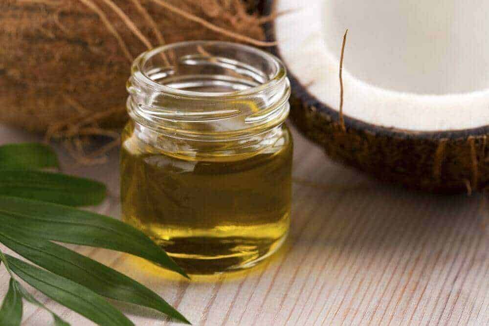 Olio di cocco biologico in barattolo trasparente, con noce di cocco e foglie di palma sullo sfondo, su superficie di legno naturale.