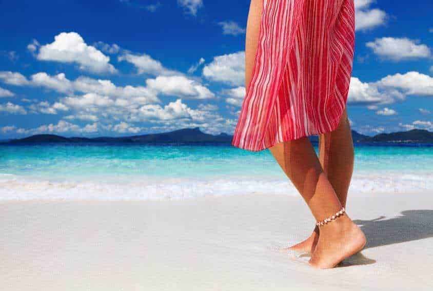 Passeggiata sulla spiaggia con abito estivo leggero e ciambella alla caviglia, ambiente marino con acque cristalline e cielo nuvoloso, ideale per relax e vacanze estive.