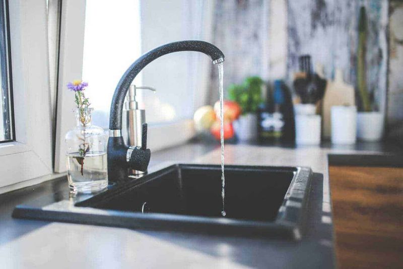 Quale lavello scegliere in cucina, singolo o doppio? 3 Lavandino da cucina con rubinetto aperto e acqua che scorre, pianta fiorita in bottiglia di vetro, sfondo con utensili e piante, ambiente domestico luminoso e accogliente.