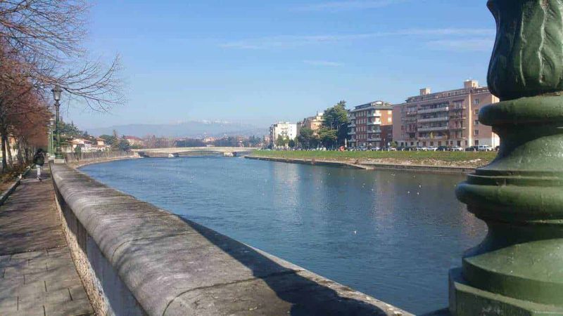 Dove si trova Verona, Meteo e cosa vedere nella Piccola Roma (2026) 8 Fiume e passeggiata urbana con edifici residenziali, in una giornata di sole, panoramica dozzina di fiume e paesaggio cittadino con acqua e cielo blu, ambiente sereno, luoghi di relax e vita cittadina.