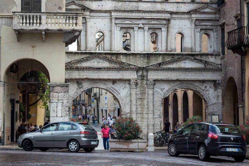 Dove si trova Verona, Meteo e cosa vedere nella Piccola Roma (2026) 11 Antica porta romana in pietra e archi storici in un centro urbano italiano, con auto parcheggiate e persone che passeggiano, rappresentando un angolo caratteristico e storico di una città italiana.