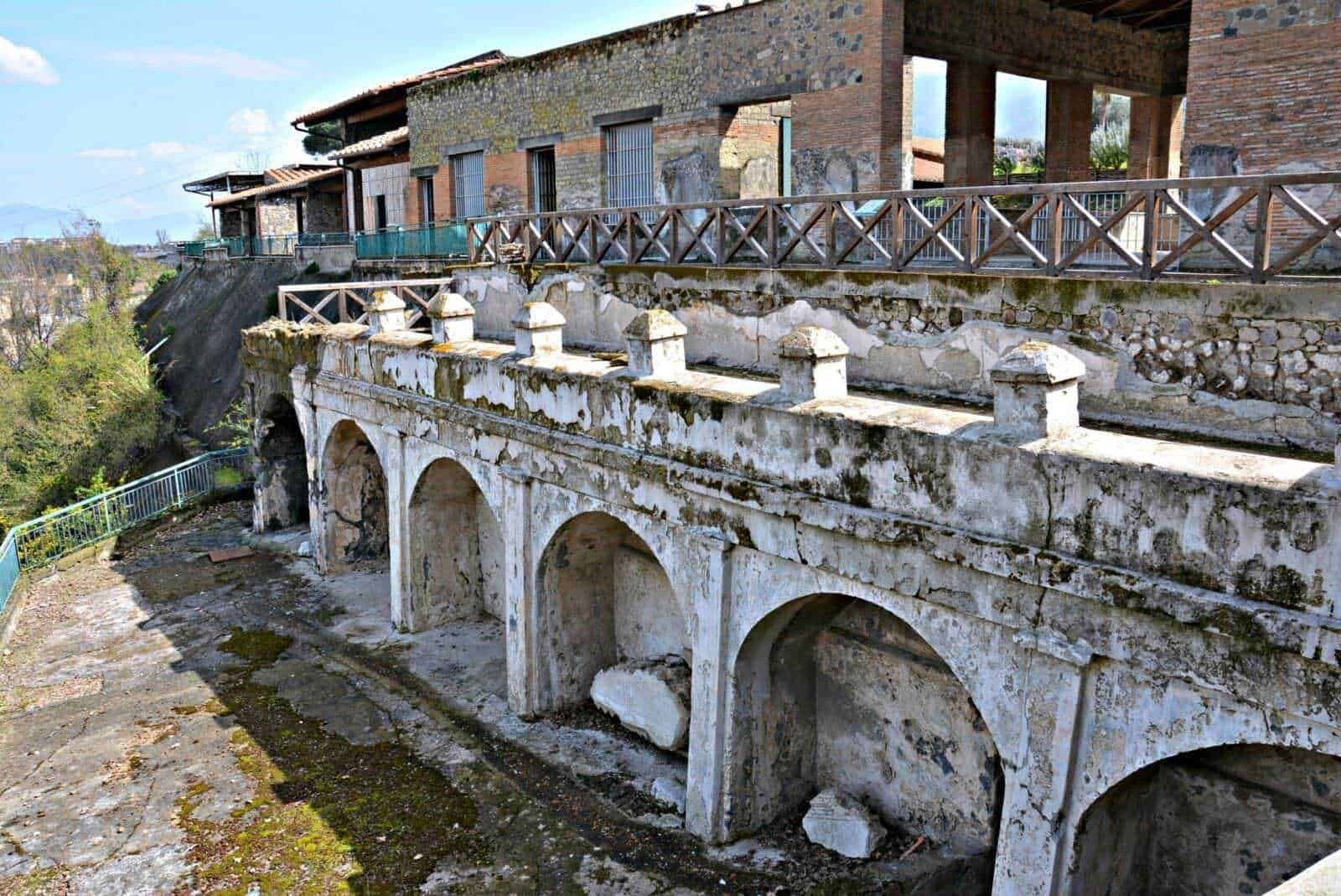 Vecchio ponte in rovina con archi in pietra e muratura, su sfondo di edifici storici, cielo blu e vegetazione circostante, resti di un passato architettonico, deteriorato ma affascinante.