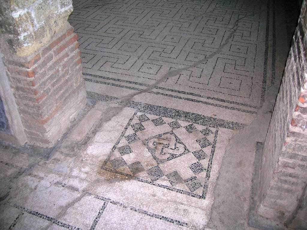 Motivo di mosaico romano con dettagli geometrici e simbolici, parzialmente scoperto e in fase di restauro, situato tra muri di mattone antico.