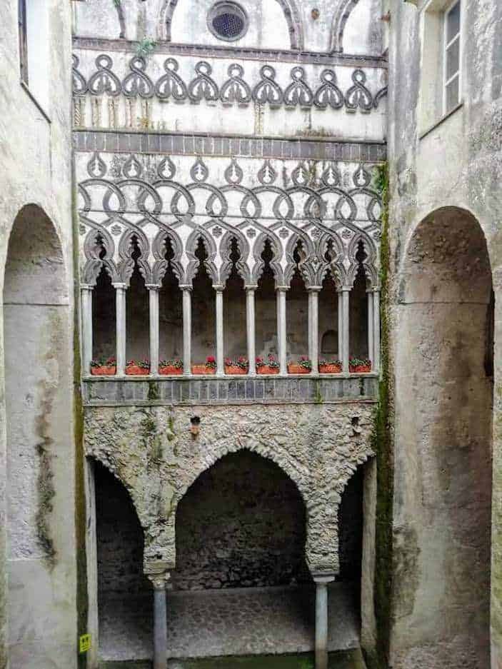 Antico cortile con archi e decorazioni in stile gotico, pareti in pietra e balcone con ringhiera decorativa e piante di fiori, ambiente storico e affascinante, esempio di architettura medievale italiana.