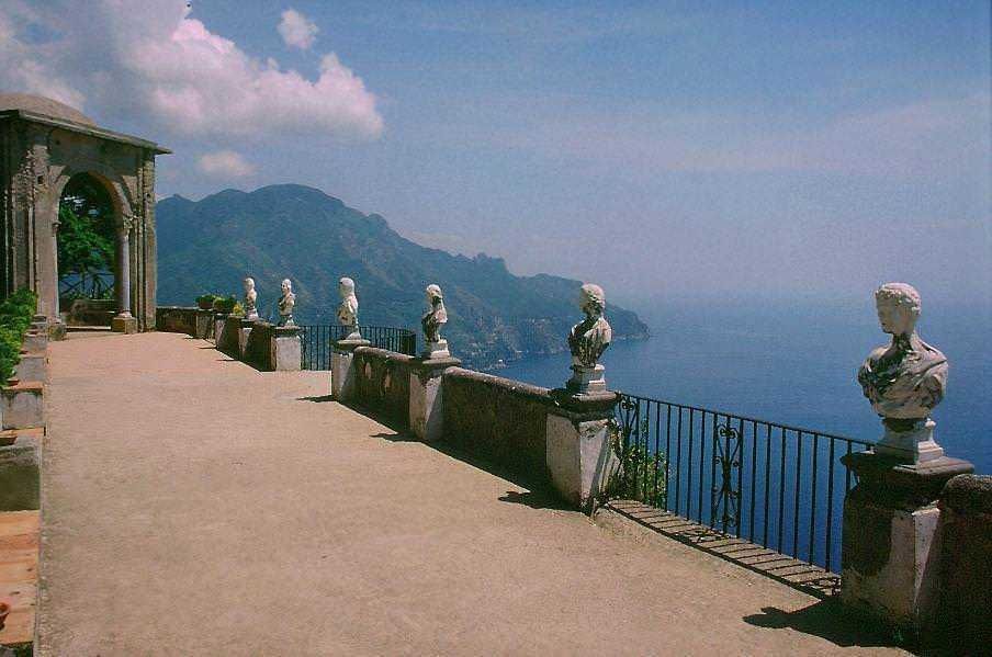 Vista panoramica sul mare e sulle scogliere con statue di busti in stile classico, sulla terrazza di un elegante edificio storico, con montagne sullo sfondo, atmosfera rilassante e suggestiva.