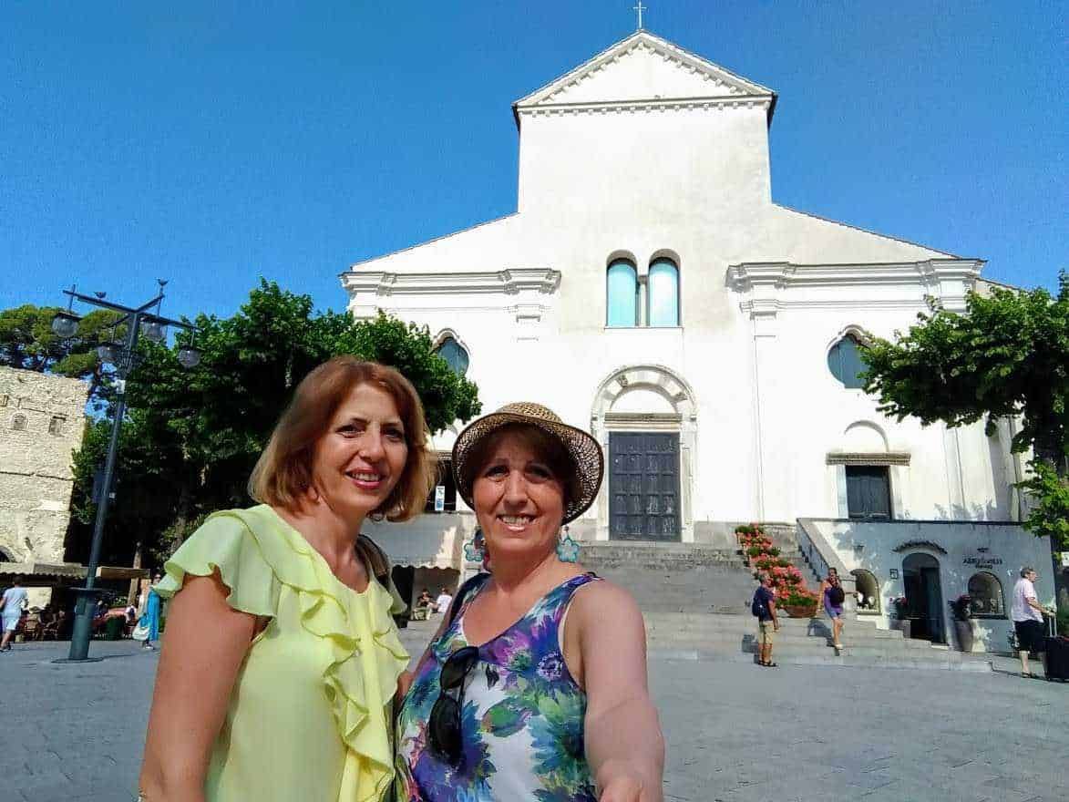 Femminili turiste in abiti estivi davanti a una chiesa bianca storica in Italia, con cielo sereno e architettura religiosa.