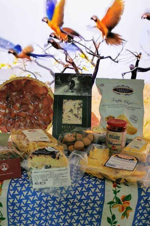 Prodotti tipici italiani online: Voli di uccelli colorati che si librano nel cielo sopra un'ampia selezione di prodotti alimentari italiani, tra cui dolci, biscotti e conserve, con uno sfondo di cielo e natura.