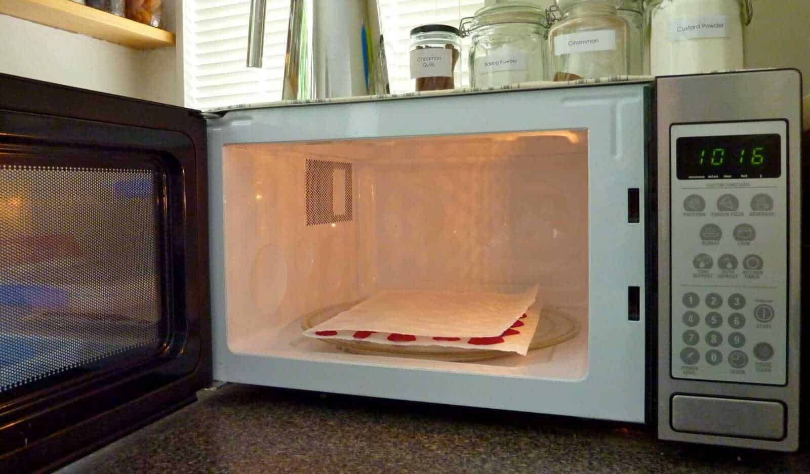 L'immagine mostra un forno a microonde con un piatto di carta e un tovagliolo all'interno, pronto per essere riscaldato o cotto. Sul ripiano superiore si vedono barattoli di spezie e aromi, come cannella e anice, in contenitori di vetro trasparente. La scena rappresenta un ambiente domestico e cucina, perfetta per contenuti legati a ricette, cucina casalinga e tecniche di riscaldamento alimentare.