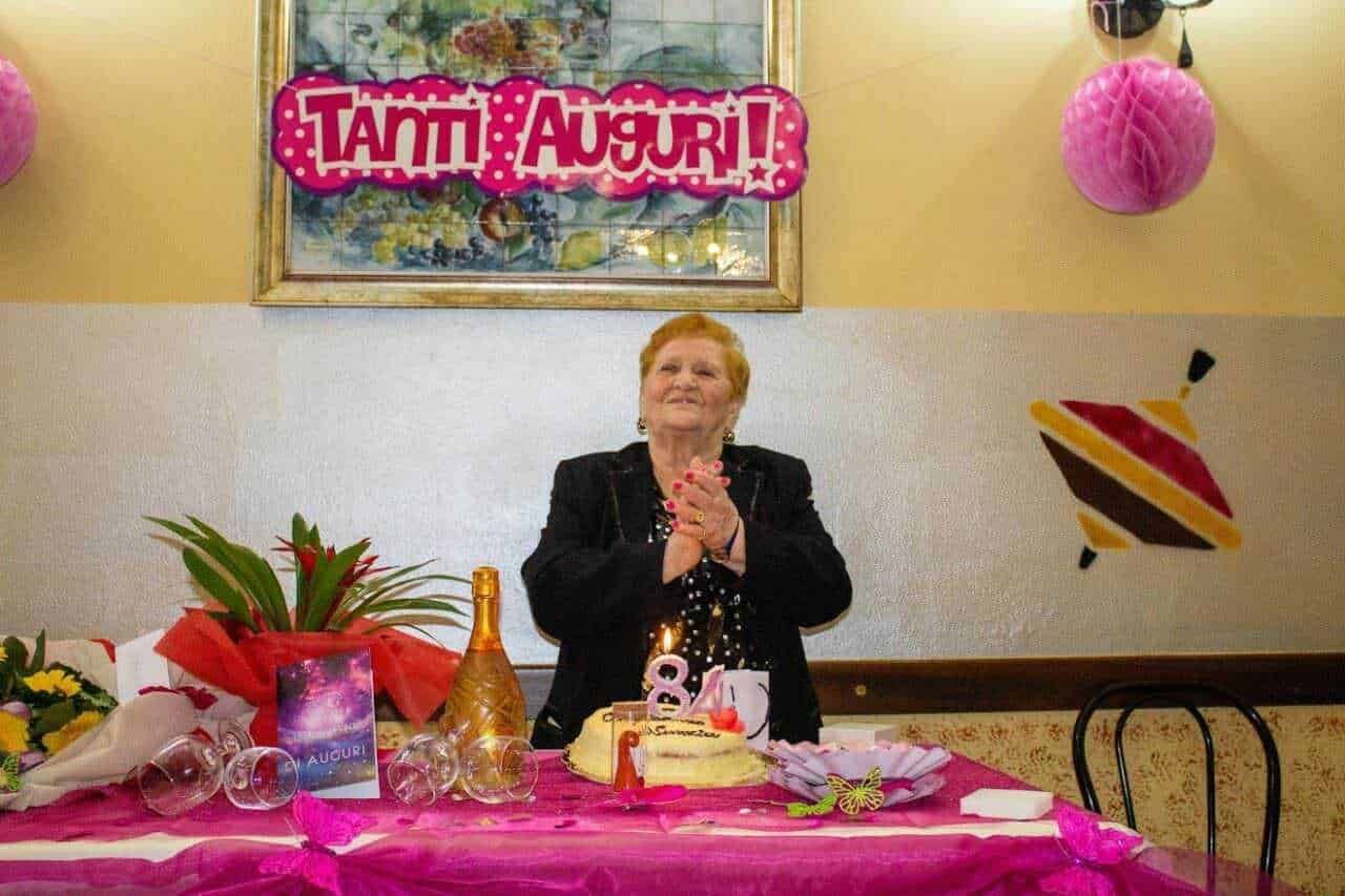 Tanti auguri a una donna anziana che festeggia il suo compleanno con torta, decorazioni rosa e regali, in un ambiente festoso per celebrare un'occasione speciale.