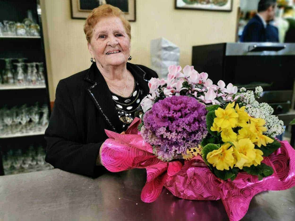Fiore di composizione floreale colorata con rosa, giallo e viola, donna anziana sorridente con abbigliamento elegante, ambiente accogliente, celebrazione speciale.
