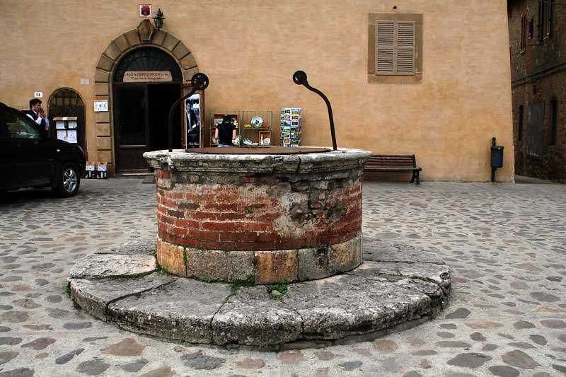 Vecchio pozzo in pietra e mattoni al centro di una piazza di città storica, con edificio antico sullo sfondo.