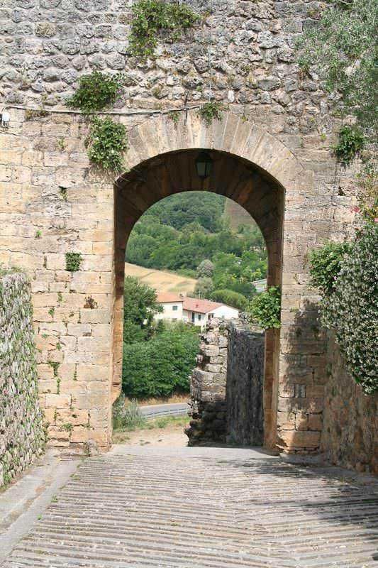 Vecchio arco in pietra con vista panoramica su un paesaggio collinare verde e case rurali, strada in ciottoli, scenario di un borgo storico italiano, atmosfera tranquilla e autentica.