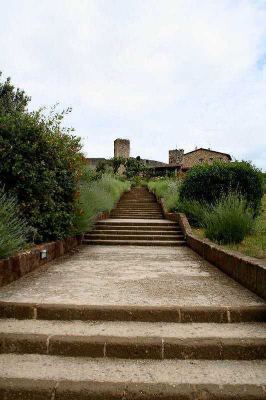 Scalinata in pietra che conduce a un borgo medievale con torri e case antiche, immerso in verde. Ideale per escursioni e passeggiate nella natura.