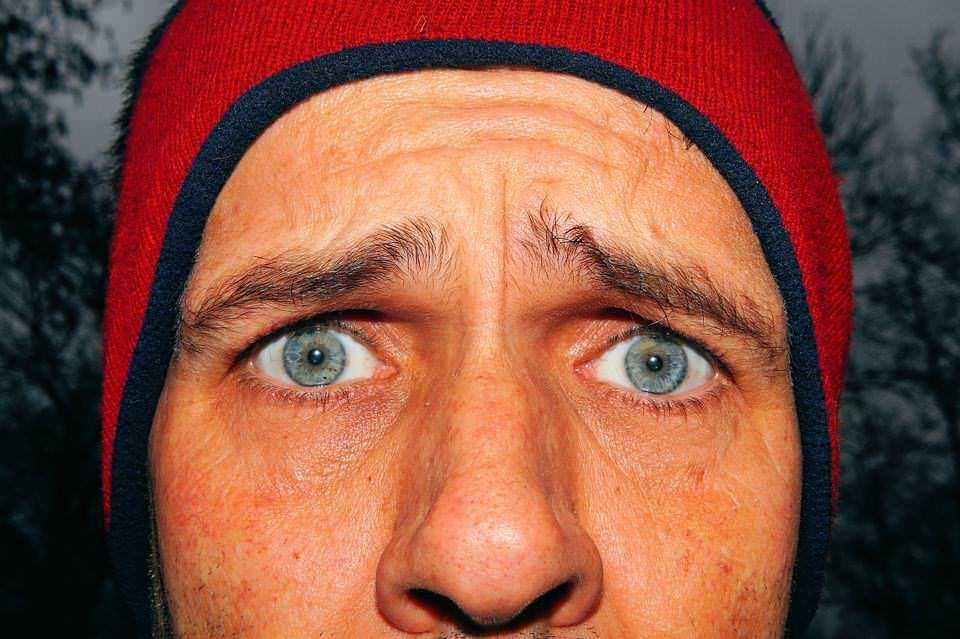 Stelle di un uomo con occhi azzurri e volto arrossato, in primo piano, indossa un cappello rosso, sfondo naturale con alberi e cielo nuvoloso, primo piano dettagliato, immagine di alta qualità, atmosfera outdoors, condizioni di luce naturale.
