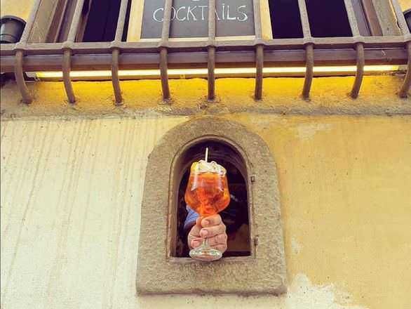 Aperitivo in un pub, mano che tiene un drink color ambra in un bicchiere con cannuccia, vista da una finestra su una strada cittadina, atmosfera accogliente e convivialità.