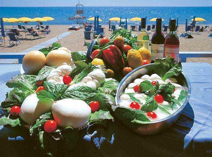 Pomodori, mozzarella e vino in una tavola sulla spiaggia, ideale per un aperitivo estivo. La scena è immersa in un ambiente marino con ombrelloni gialli e lettini sulla sabbia.